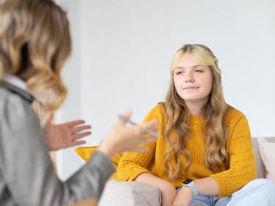A girl sits and listens to a woman gesturing with her hands and holding a pen